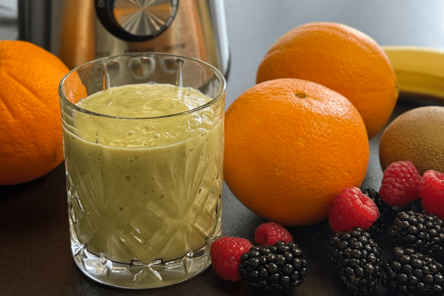 Grüner Anti-Kater-Smoothie in einem Glas vor einem Standmixer, dekoriert mit frischem Obst, das um das Glas angeordnet ist.