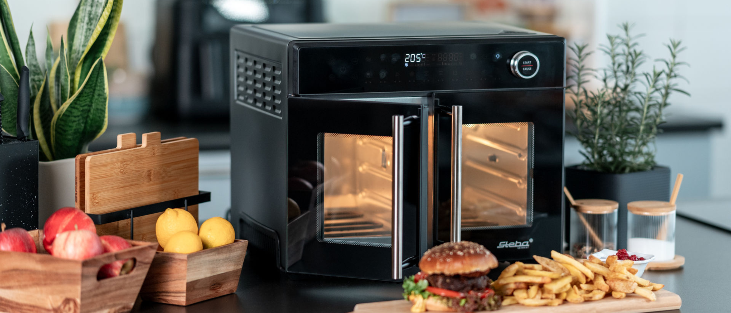 Airfry-Grillbackofen AF 24 auf einer Küchentheke angeschaltet, mit einer geöffneten Tür. Vor dem Gerät befindet sich ein Holzbrett auf dem Ein Hamburger sowie Pommes angerichtet sind.