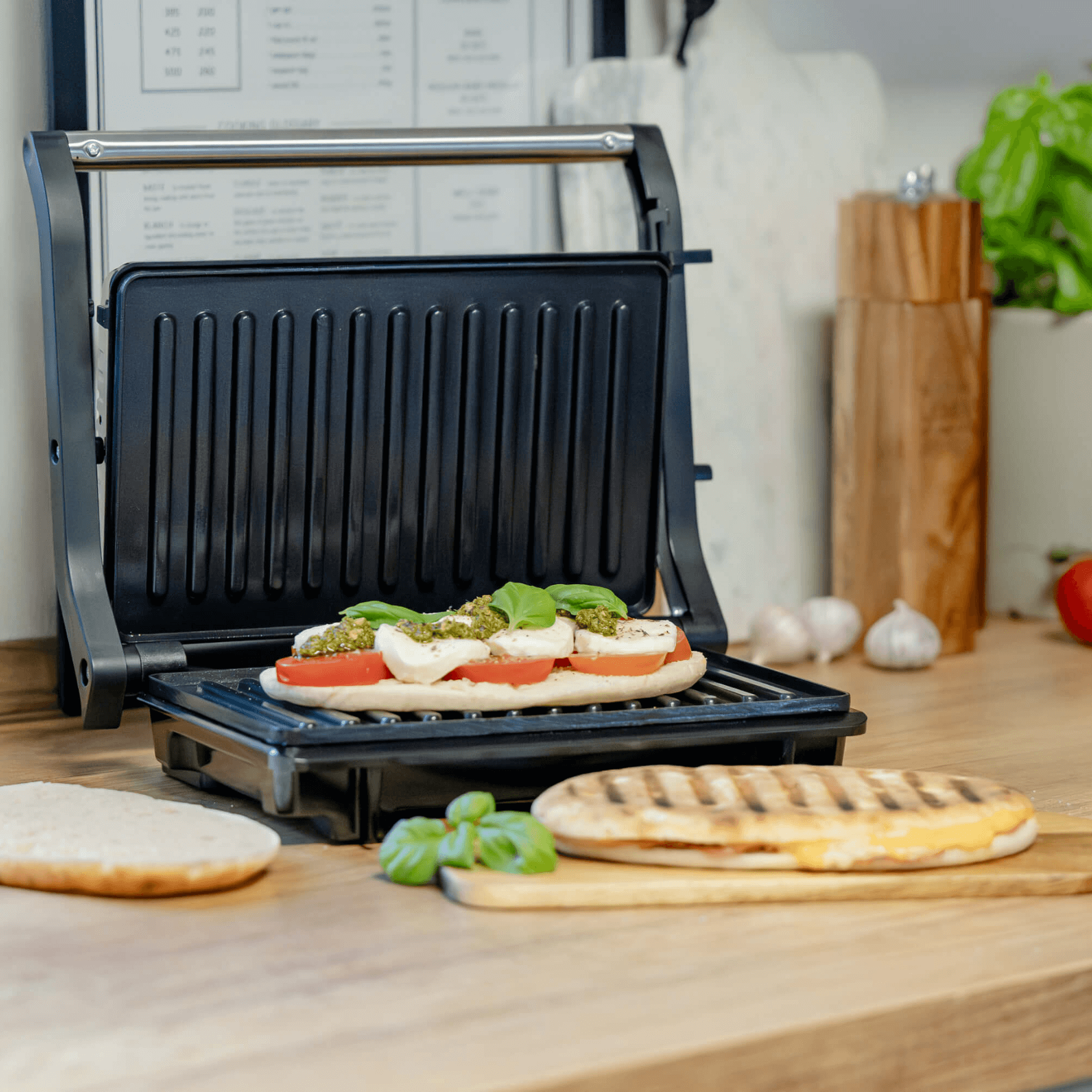 Kontaktgrill von Steba mit geöffnetem Deckel, belegt mit Tomaten, Mozzarella und Basilikum. Holzunterlagen und frische Zutaten sichtbar.