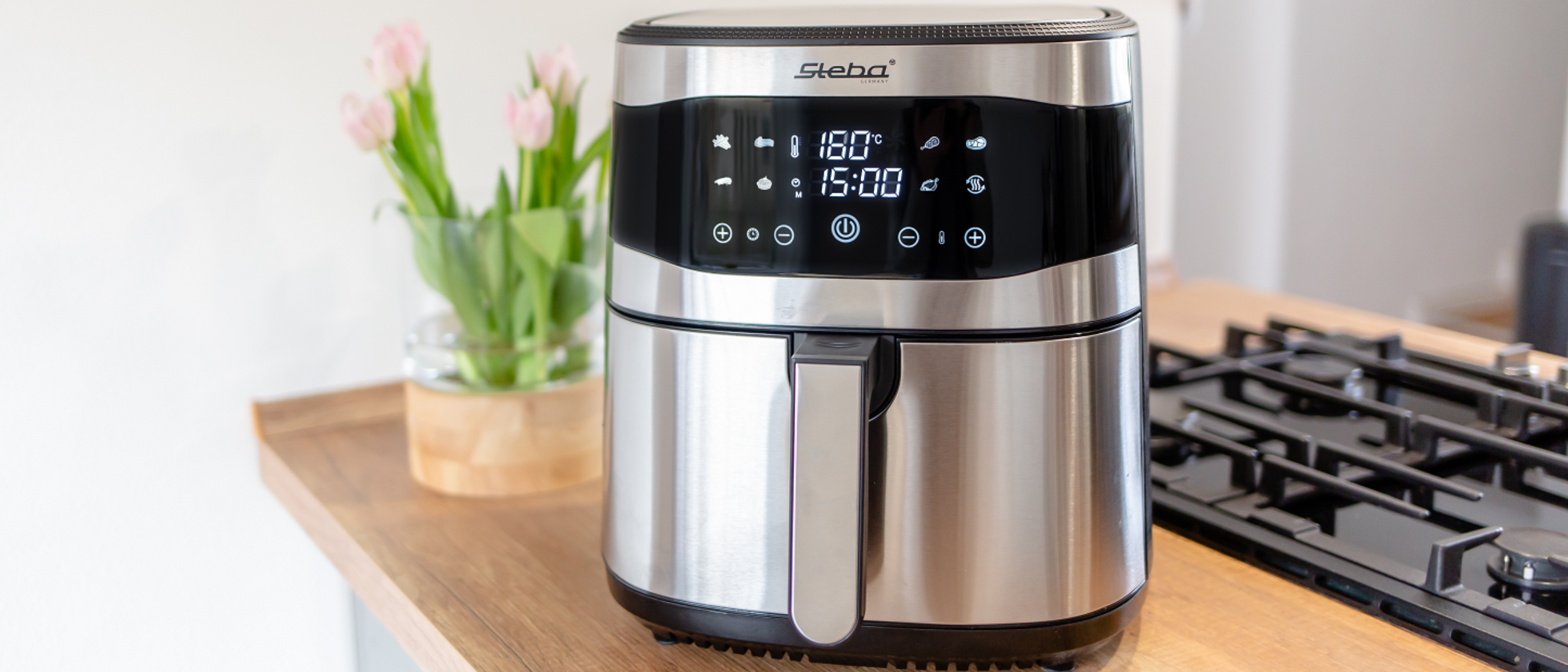 Heißluftfritteuse HF 8000 auf einer Küchenthenke neben einem Herd. Das Gerät ist geschlossen und angeschaltet. Das digitale Display zeigt Uhrzeit sowie Grad an.