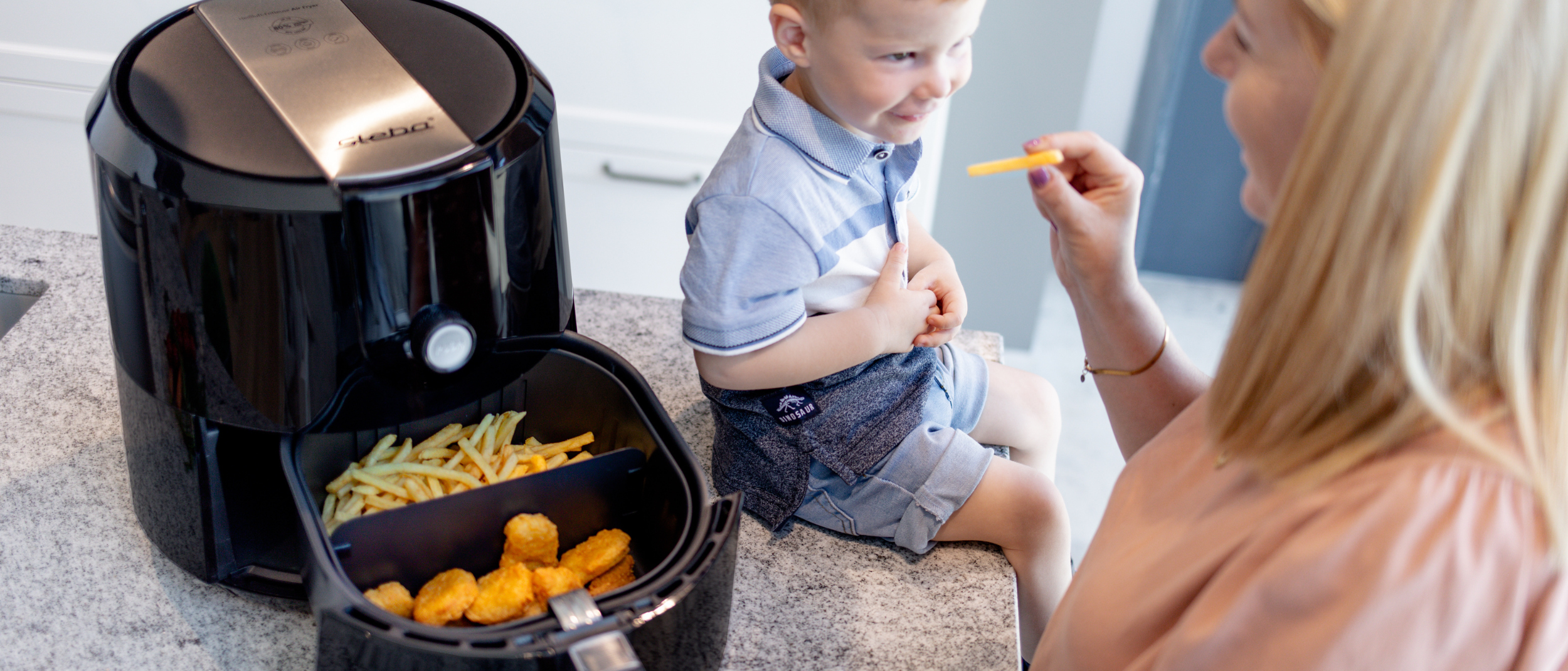 Heißluftfritteue HF 5000 geöffnet, mit einer Trennwand im Frittierkorb, die Pommes und Nuggets trennt. Die Heißluftfritteuse steht auf einer Küchentheke, auf der ein Kleinkind sitzt, das gerade eine Pommes von einer Frau bekommt.