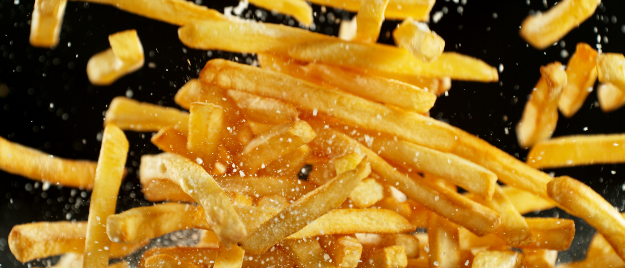 Mehrere goldgelb frittierte Pommes, die in der Luft schweben, während Salz darüber gestreut wird