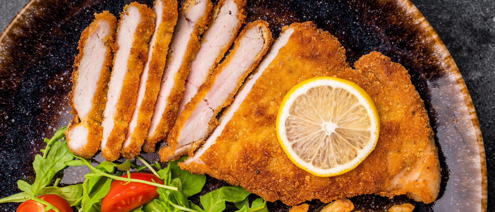 Ein paniertes Schnitzel auf einem Teller, angerichtet auf einem Salatbett mit einer Zitronenscheibe darauf.