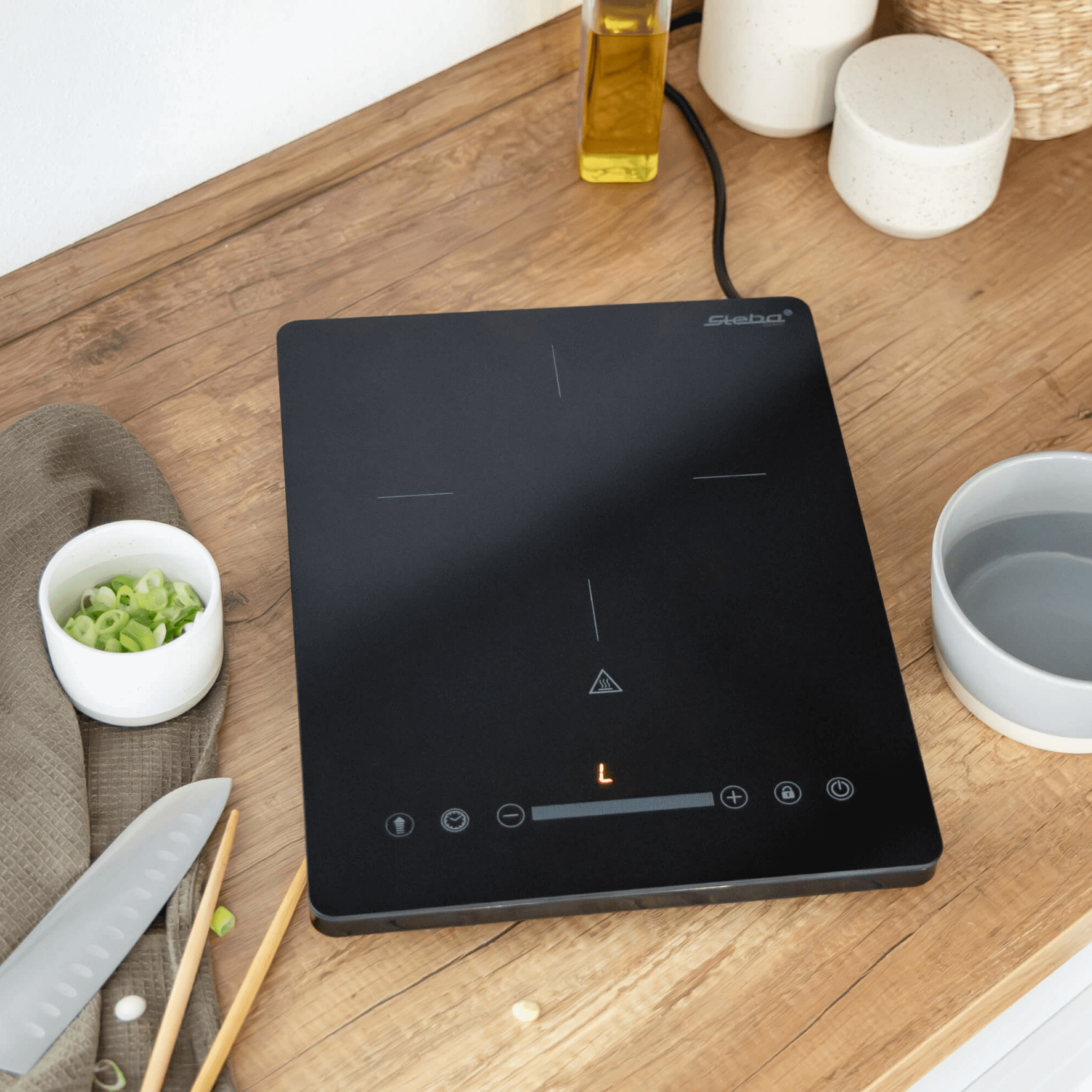 Induktionskochfeld von Steba, flache schwarze Oberfläche mit Touch-Bedienelementen, umgeben von Küchenutensilien auf einem Holztisch.