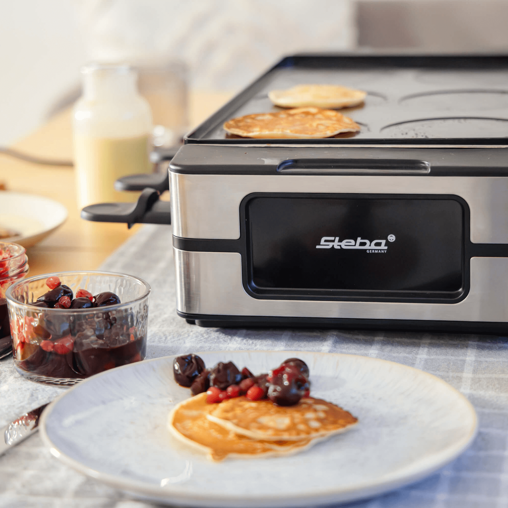 Steba Raclette-Grill RC180 mit Pfannkuchen auf der Grillplatte, serviert mit Marmelade und Beeren. Edelstahl-Gehäuse.