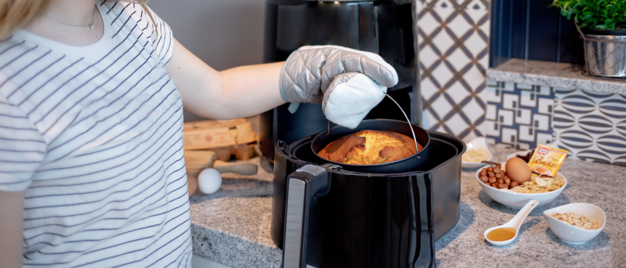 Heißluftfritteuse HF 5000 auf einem Küchentresen, umgeben von Backutensilien. Der Korb ist offen und daraus wird eine Backform mit frischen Kuchen herausgehoben.